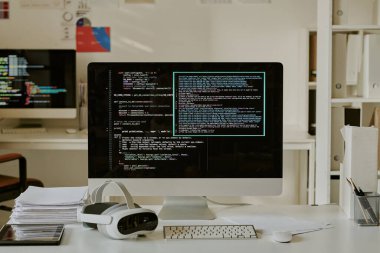 Bilgisayar şirketinin ofisindeki bilgisayarın yanında devasa monitör gösterim kodları, kağıt yığını, tablet ve VR kulaklığı olan orta boy çalışma masası.