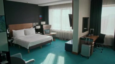 Geniş otel odasının içinde çift kişilik yatak, aynalı masa, kumsal zemininde koltuk ve bavul, pencereden içeri güneş ışığı akışı.