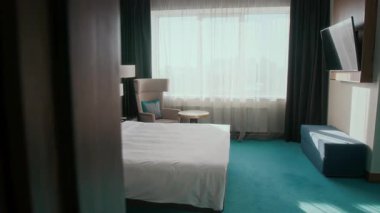 Modern otel odasının içinde çift kişilik yatak, yastıklı koltuk, yerde lambalar ve duvarda televizyon, büyük pencerelerden süzülen gün ışığı.