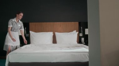 Klasik üniformalı kadın hizmetli, misafir gelmeden önce otel odasında yatağını toplarken beyaz battaniye ve yastıkları dikkatlice düzenliyor.