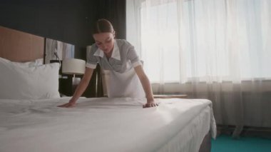 Üniformalı kadın hizmetçi otel odasında güneşli çarşafları düzleştiriyor.