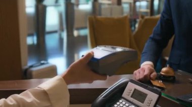 Otelin lobisindeki ön masada duran erkek misafirin yakın plan resmi. Kadın resepsiyonistin elindeki banka kartına dokunarak ulaşılamaz bir ödeme yapıyor.