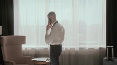 İş adamı otel odasında yürüyor, belgeleri okuyor ve telefon görüşmeleri sırasında pencereden bakıyor.