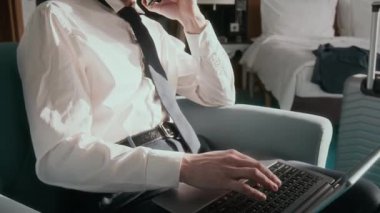 Formalite icabı koltukta oturan, cep telefonuyla konuşan ve otel odasında iş gezisi sırasında dizüstü bilgisayarda yazan bir adamı çek.