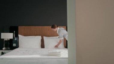 Üniformalı kadın hizmetçi yatağını topluyor, sonra yeni havlular alıp otel odasında geziyor ve misafirler için hazırlanıyor.