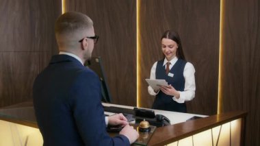 Gülümseyen kadın resepsiyonist, üst sınıf otel resepsiyonunda kontrol işlemini tamamlarken erkek müşteriden dijital tablet imzalamasını istiyor.
