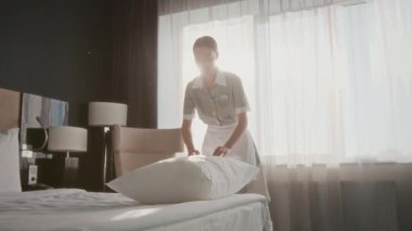 Kadın otel hizmetçisi üniformalı. Misafir odasında yastığı kabartıyor. Parlak güneş ışığı pencereden içeri süzülüyor.