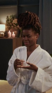 Uzun örgülü, cüppeli, gülümseyen ve kaplıcada kameraya poz veren Afro-Amerikan bir kadının dikey portresi.