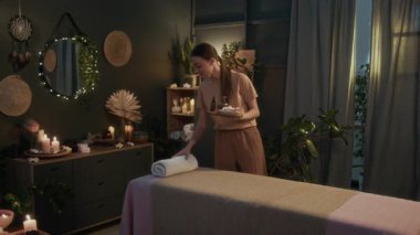 Yavaş çekim, masaj için hazırlanan kadın uzmanlar, sakin spa odasında, ahşap mobilyalar, bitkiler ve mumlarla müşterilerini bekliyorlar.