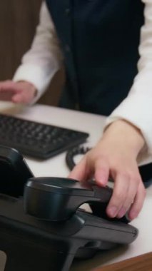 Kadın otel resepsiyonistinin bilgisayarı kullanırken, telefonunu açarken ve ön masada çalışırken konuşurken dikey eğim görüntüsü.