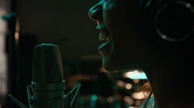Kulaklıklı Afro-Amerikalı genç bir kızın profesyonel mikrofona şarkı söylerken duygusal şarkılar kaydetmesi, loş bir müzik stüdyosunda.