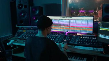 Genç erkek ses mühendisi, kontrol odasında dijital karıştırma konsolu kullanarak genç grupla müzik kaydı yaparken kulaklık takıyor.