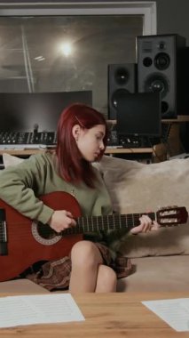Akustik gitarla şarkı söyleyen beyaz bir kızın dikey görüntüsü, arka planda oturan Afro Amerikalı bir çocuk.