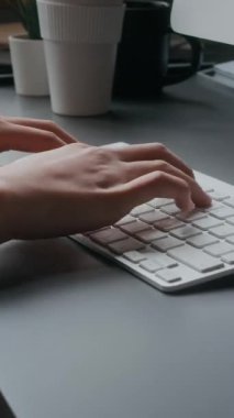 Asyalı genç bir kadının ofis masasındaki kablosuz bilgisayar klavyesine tekrar kullanılabilir kahve fincanı ve bitkilerle yazı yazarken dikey eğim görüntüsü.