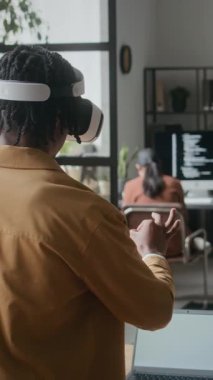 VR kulaklık takan Afro-Amerikan bir adamın dikey çekimi, ofiste duruyor ve yeni uygulamayı test ederken havadaki sanal elementlerle etkileşime geçiyor.