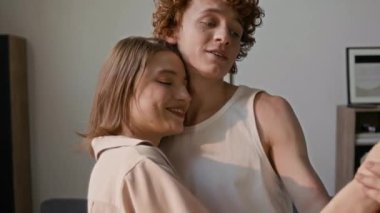 Genç ve sevecen bir çift müzik eşliğinde dans ediyor ve evlerinin oturma odasında mutlu bir şekilde gülümsüyor.