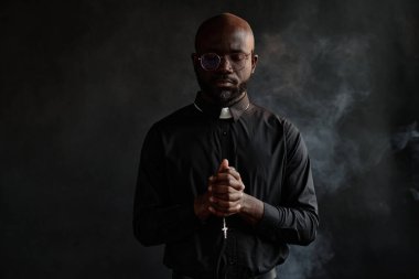 Siyah cüppeli genç Afrikalı Amerikalı rahibin sessizce dua ederken haçla haç taşıdığı orta boy bir fotoğraf. Karanlık odada tütsüsü havada süzülüyor.