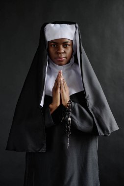 Siyah beyaz giysili Afro-Amerikan rahibenin dikey portresi sabit duruyor ve tespihi elinde tutuyor.