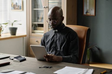Siyah cüppeli genç Afrikalı Amerikalı rahibin minimalist ofiste ahşap mobilyalarla tablet kullanırken rahat deri koltukta oturduğu orta boy bir fotoğraf.