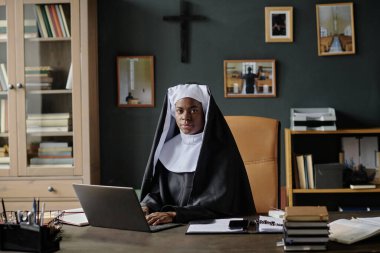 Üzerinde dizüstü bilgisayar, not defteri ve ofis malzemeleri olan ahşap masada otururken Afrikalı Amerikalı rahibenin kameraya baktığı geniş açı.
