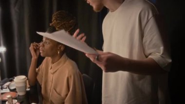 Meslektaşı makyajını yaparken ve loş ışıklandırılmış soyunma odasında onu dinlerken, erkek stand-up sanatçısının performansını prova ederken.