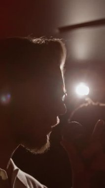 Sakallı ve gözlüklü genç adamın loş ışıkta mikrofona konuştuğu dikey yakın çekim.