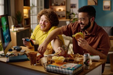 İki arkadaş video oyunlarına odaklanırken abur cubur ve içkinin tadını çıkarıyor. Masanın üzerine çeşitli atıştırmalıklar ve içecekler serpiştirilmiş modern bir daire.