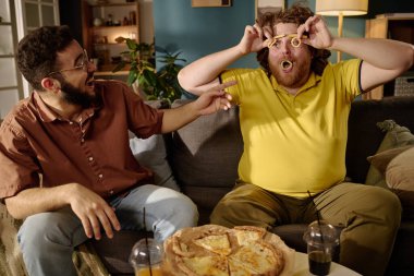 İki arkadaş keyifli bir anın tadını çıkarıyorlar. Oturma odasında gülümsüyorlar ve önlerinde lezzetli bir pizza ile eğleniyorlar.