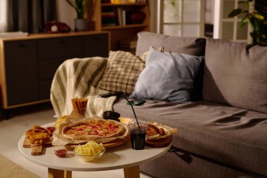 Sıcak ve cezbedici oturma odası sahnesinde lezzetli bir pizza, patates kızartması ve ferahlatıcı bir içecek, rahat bir kanepe ve yumuşak yastıklı rahat atmosferi vurgulayan bir masa seti.