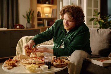 Kıvırcık saçlı bir adam kanepeye oturmuş, kitaplarla çevrili sıcak oturma odasında pizza ve atıştırmalıkların tadını çıkarıyor. Öndeki ahşap masa lezzetli yiyeceklerle dolu.