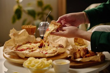 Erimiş peynirli peynirli bir dilim pizzayı çeken bir elin yakın çekimi. Etrafı yumuşak bir içecek, waffle külahı ve iyi hazırlanmış bir yemek masasında limon dilimleri ile çevrili.