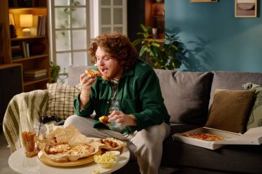 Genç adam kanepede oturuyor, rahat ve rahat bir ortamda bir dilim pizzanın tadını çıkarıyor. Masaya yerleştirilmiş pizza kutuları, atıştırmalıklar ve içecekler de dahil olmak üzere çeşitli şeyler.