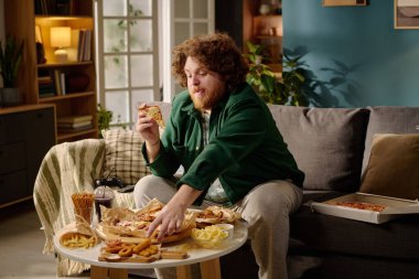 Kanepede otururken pizza ve abur cubur yiyen kıvırcık saçlı genç adam. Oda sıcacık dekorlar, bitkiler ve raflarla dolu. Sıcak ve davetkar bir ortam yaratıyor.