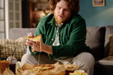 Oturma odasında rahat koltukta oturan bir dilim peynirli pizza tutan ve pizzadan erimiş peynir alan bir adam. İçecekler ve atıştırmalıklar yanında rahat bir yemek atmosferi yaratıyor.