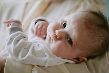 Bebeğin ebeveynlerinin kollarında sevgi ile sarılırken gözleri açık dinlenmesi. Atmosfer sıcaklık ve güvenlik hissi yayıyor.