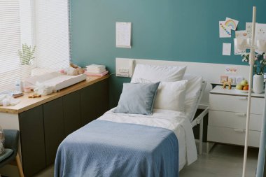 Pastel dekorlu rahat bir yatak odası, mavi battaniyeli temiz bir yatak, duvarları süsleyen çeşitli dekorasyonlar ve komodinli. Yumuşak doğal ışık panjurlardan içeri giriyor