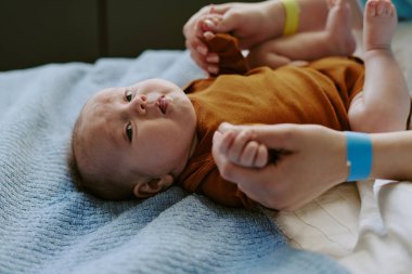 Bebeğin kahverengi tulum giyip, yatakta uzanırken anne ve babasının parmaklarını kavradığı yakın plan. Çocuk ve ebeveyn arasında hassas bir an.