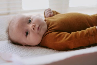 Bebek portresi, rahat kahverengi uzun kollu bir elbise içinde yatağa uzanmış, kameraya tuhaf bir şekilde bakıyor, arka plandaki pencereden süzülen yumuşak doğal ışık.