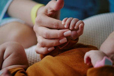 Yetişkin ellerinin, yeni doğan bebeğin minik elini tutarak hassas bir bağ ve nazik bir bakım göstermesi. Bebek kahverengi bir battaniyeyle kundaklanmış, sıcaklık ve koruma çağrıştırıyor.