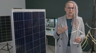 Alternatif enerji çözümleri sunan bir kadın, ofiste büyük bir güneş panelinin önünde minyatür rüzgar türbini tutuyor.