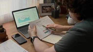 Kağıda grafikle bakan ve ekrandaki grafiklerle karşılaştıran uzaktan çalışan işçinin omuz manzarası