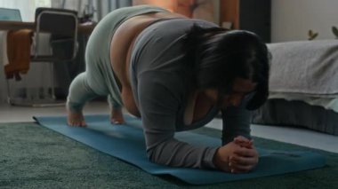 Spor kıyafetleri giymiş kıvrımlı genç bir kadın tahtada duruyor ve evde bir dakika tutmaya çalışıyor.