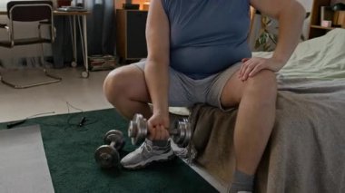 Tıknaz, beyaz bir adam kenarda oturuyor ve dambılla biseps yapıyor.