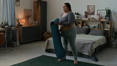 Latin bir kadın koyu yeşil halıda spor paspası açıyor. Evde yoga yapmaya başlıyor.