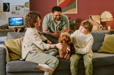 Kafkasyalı kadın, beyaz çocuk ve Orta Doğulu adam kanepeye oturmuş küçük kahverengi köpekle etkileşime geçiyorlar. Yetişkinler gülümserken ve evdeki ilk evcil hayvanı izlerken evcil hayvanı nazikçe okşuyorlar.