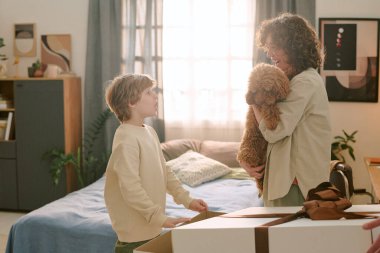 Beyaz bir kadın kucağında küçük bir köpek, yatak odasında kutusunu açan beyaz bir çocuğa gülümserken ilk evcil hayvanı aile üyeleri arasında görünür bir heyecan ve etkileşimle yakalıyor.