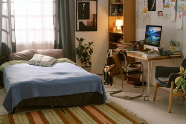 Yatak odasındaki rahat sahne mavi battaniyeli yatağı, bilgisayar monitörlü masayı, kitapları ve sırt çantasını, kapalı alanda bitkileri ve duvar resimlerini gösteriyor.