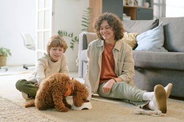 Beyaz bir kadın yerde oturmuş gülümserken beyaz bir çocuk küçük kahverengi bir köpeği okşuyor. Oturma odasında klozetten su içiyor. Ev tecrübesindeki ilk evcil hayvanı canlandırıyor.