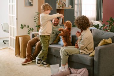 Kafkas çocuk küçük kahverengi köpekle oynarken ailesi kanepede oturmuş oturma odasındaki etkileşimi izliyor.