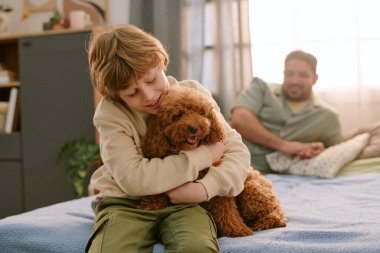Beyaz çocuk, orta yaşlı beyaz adam arka planda izlerken tüylü kahverengi köpeğe sarılıyor. Evdeki ilk evcil hayvanla neşe dolu bir bağ kurduğu anı yakalıyor.